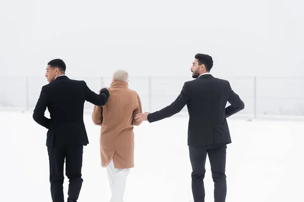 Back view of mature businesswoman walking across snowy field under protection of interracial bodyguards in black suits — Stock Photo