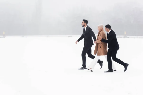 Side view of senior businesswoman walking across snowy field under protection of interracial bodyguards — Stock Photo