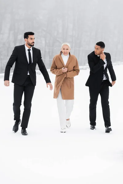 Full length view of interracial bodyguards in black suits escorting mature businesswoman on snowy field — Stock Photo