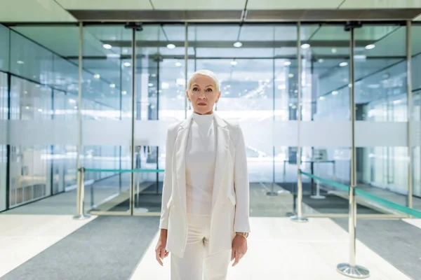 Seniorin im weißen Anzug steht in Hotellobby in der Nähe des Eingangs — Stockfoto