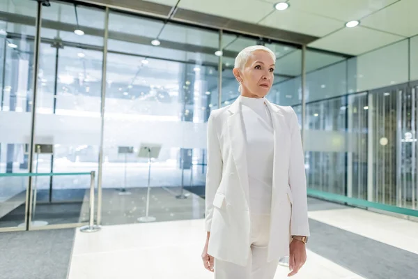 Reife Geschäftsfrau im weißen Anzug steht im Saal eines modernen Hotels — Stockfoto