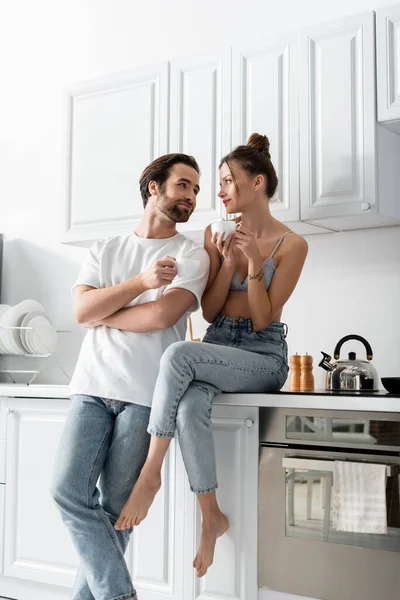 Fröhliche Frau mit Tätowierung hält Tasse Kaffee in der Hand und sitzt neben Freund in der Küche — Stockfoto