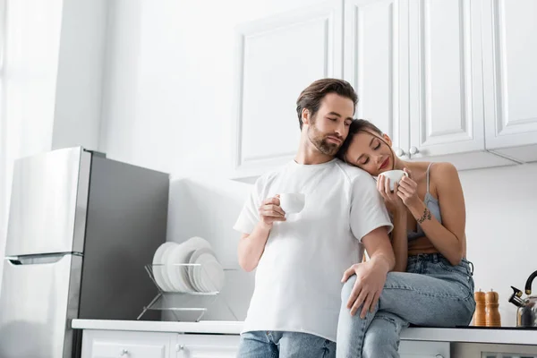 Young woman with tattoo holding cup of coffee and leaning on shoulder of boyfriend in kitchen — Stock Photo