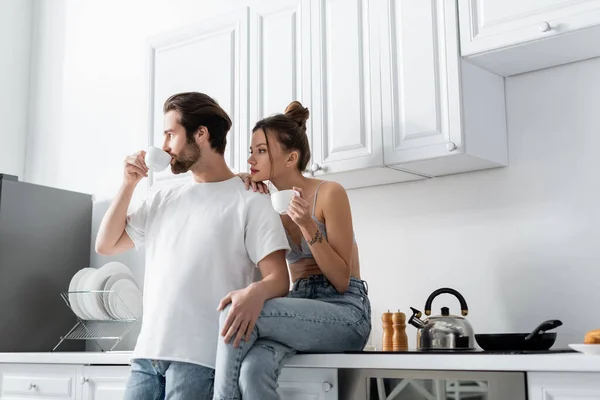 Junge Frau mit Tätowierung hält Tasse und sitzt neben Freund und trinkt Kaffee in Küche — Stockfoto