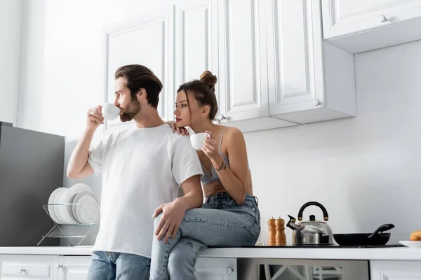 Joven mujer con tatuaje celebración taza y sentado cerca novio beber café en cocina - foto de stock