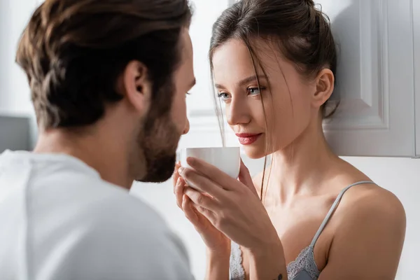 Mujer joven en sujetador sosteniendo la taza y mirando borrosa novio barbudo - foto de stock