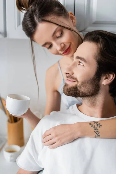 Tattooed young woman in bra holding cup and hugging pleased boyfriend — Stock Photo