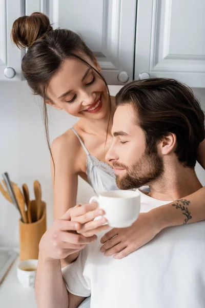 Mujer joven tatuada en sujetador sonriendo y sosteniendo la taza mientras abraza novio barbudo - foto de stock