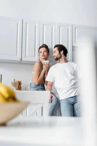 Tatoué jeune femme en soutien-gorge tenant tasse près de copain barbu dans la cuisine — Photo de stock