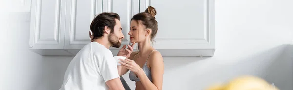 Barbudo homem seduzindo jovem mulher no sutiã com copo na cozinha, banner — Fotografia de Stock