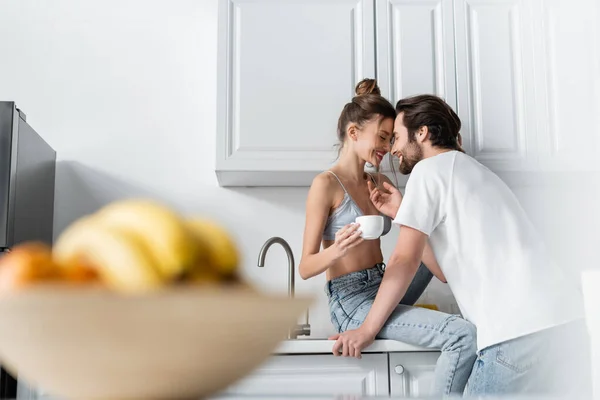 Счастливая молодая женщина в бюстгальтере держа чашку и улыбаясь с парнем на кухне — стоковое фото