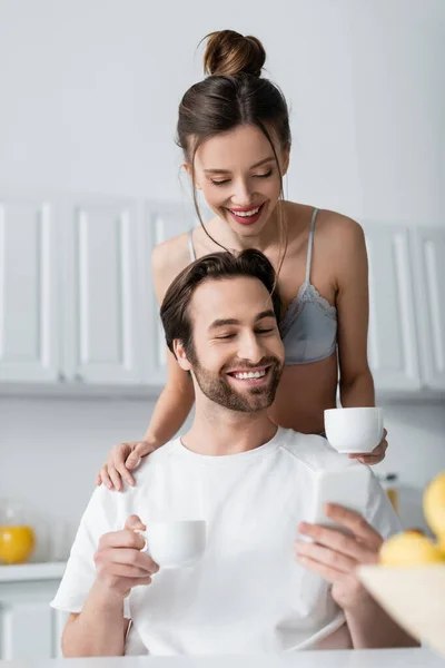 Hombre barbudo feliz usando teléfono inteligente cerca de mujer alegre en sujetador celebración taza - foto de stock