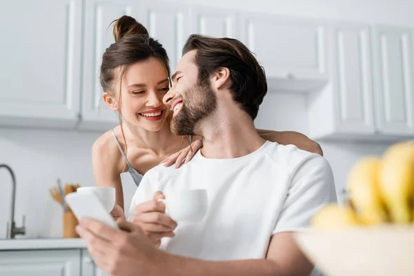 Homem barbudo feliz usando smartphone perto de mulher sorridente em sutiã segurando copo — Fotografia de Stock