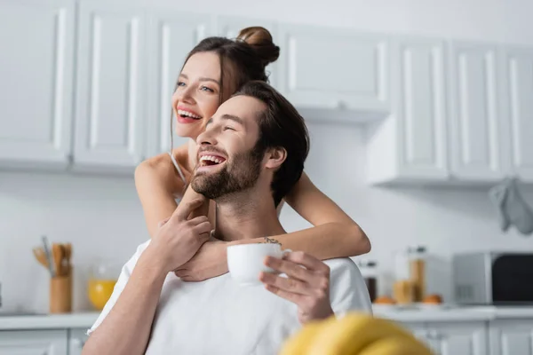 Joven mujer tatuada abrazando sonriente hombre en la cocina - foto de stock