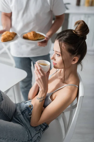 Tätowierte Frau hält Tasse neben verschwommenem Mann mit Croissants — Stockfoto
