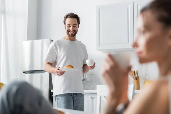 Happy bearded man holding plate with croissant and cup near blurred woman on foreground — Stock Photo