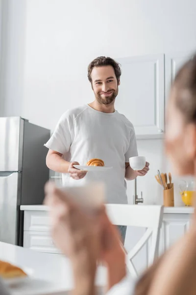 Hombre alegre sosteniendo plato con croissant y taza cerca de la mujer borrosa en primer plano - foto de stock