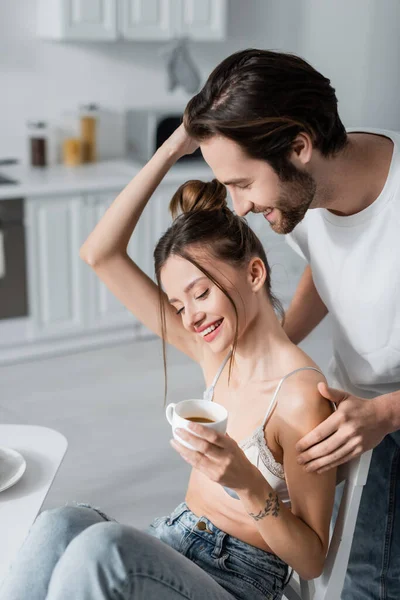 Happy tattooed woman in bra holding cup while hugging positive man — Stock Photo