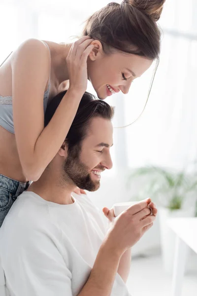 Vista lateral de la mujer alegre en sujetador de seda de pie cerca de novio con taza - foto de stock