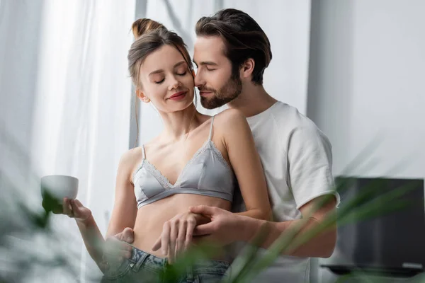 Homme barbu étreignant jeune femme en soutien-gorge et jeans avec tasse — Photo de stock