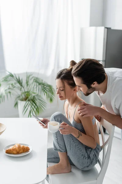 Jeune femme en soutien-gorge et jeans à l'aide d'un smartphone et tenant tasse près de petit ami dans la cuisine — Photo de stock