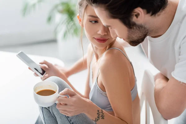 Tattooed young woman in bra and jeans using smartphone and holding cup near boyfriend in kitchen — Stock Photo