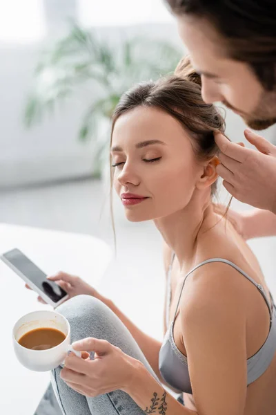 Homem desfocado ajustando o cabelo da jovem mulher em sutiã e jeans segurando smartphone e xícara — Fotografia de Stock