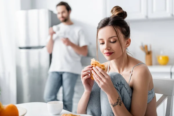 Jeune femme manger croissant près flou homme sur fond — Photo de stock