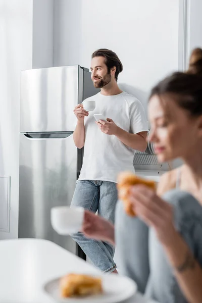 Cheerful bearded man holding smartphone and cup near blurred woman on foreground — Stock Photo