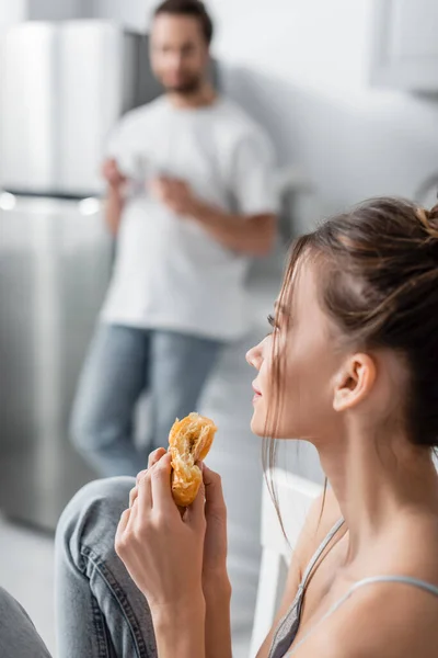 Sonhador jovem mulher segurando croissant fresco perto namorado borrado no fundo — Fotografia de Stock