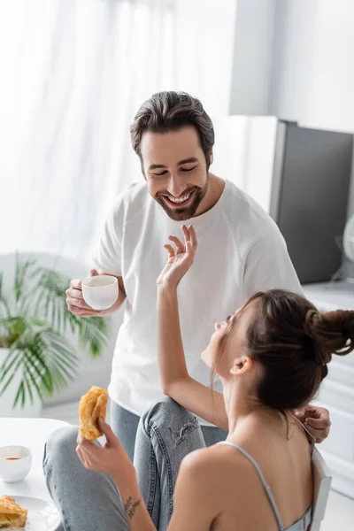 Joven mujer sosteniendo croissant fresco y llegar feliz novio con taza - foto de stock