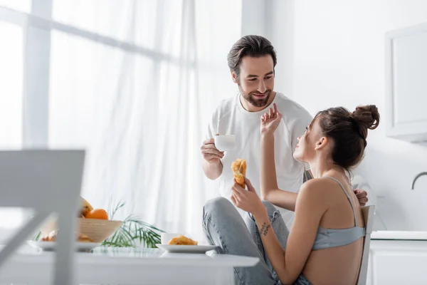 Tattooed young woman holding fresh croissant and reaching happy boyfriend with cup — Stock Photo