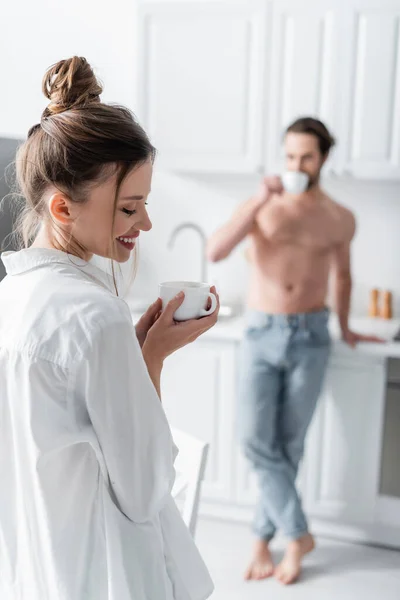 Jovem mulher em camisa branca segurando xícara de café perto de homem desfocado e sem camisa no fundo — Fotografia de Stock