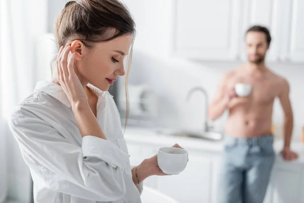 Sensual jovem mulher em camisa branca segurando xícara de café perto de homem desfocado e sem camisa no fundo — Fotografia de Stock