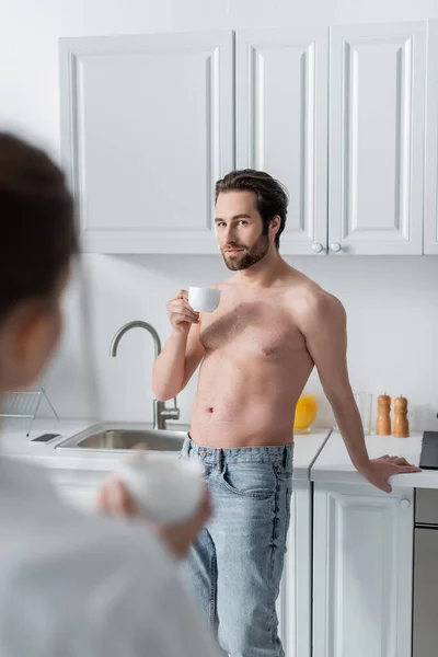 Hombre sin camisa sosteniendo la taza y mirando a la mujer borrosa - foto de stock