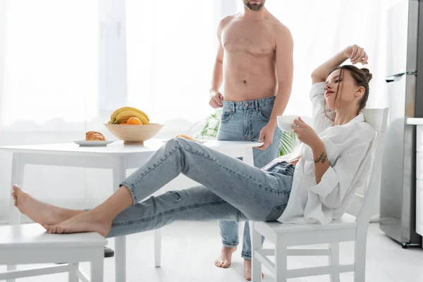 Barbudo hombre en jeans de pie cerca de la mujer joven con tatuaje en la cocina — Stock Photo
