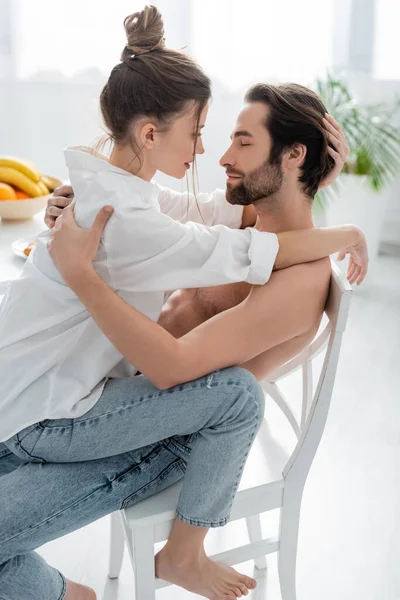 Vista lateral da mulher apaixonada sentado no homem barbudo e sem camisa na cozinha — Fotografia de Stock