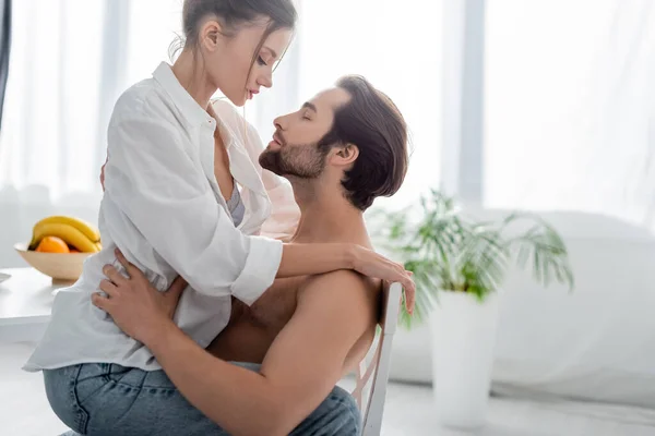 Vista lateral de la mujer seductora sentado en el hombre barbudo y sin camisa en la cocina - foto de stock