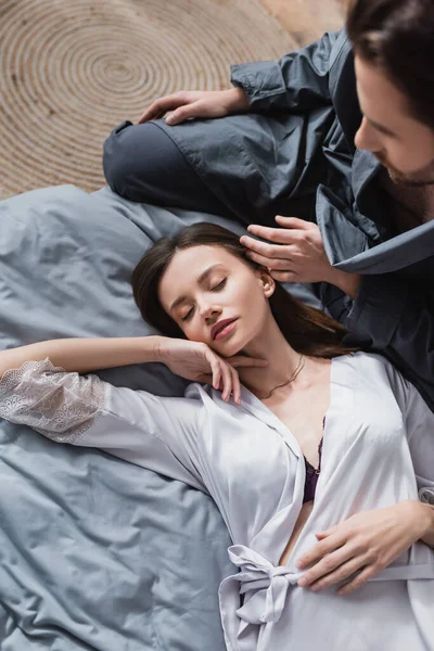Вид сверху мужчины, трогающего волосы молодой женщины в шелковом халате, лежащей на кровати — стоковое фото