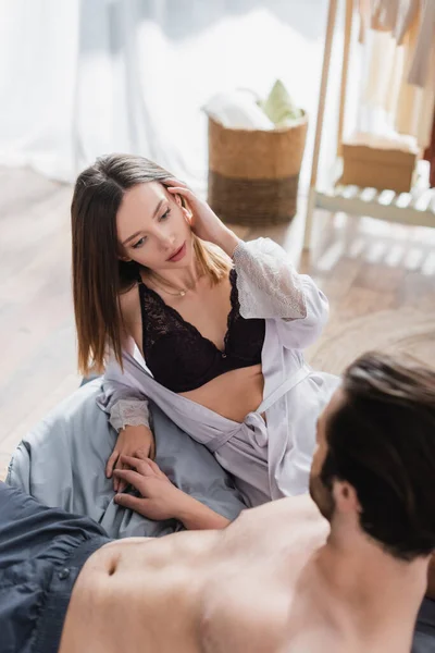 Young woman in silk robe and black bra looking at blurred shirtless man — Stock Photo