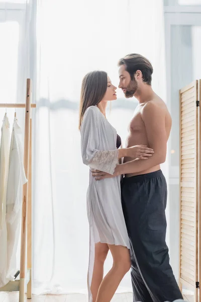 Vista lateral de la joven mujer en bata de seda abrazándose con el hombre sin camisa - foto de stock