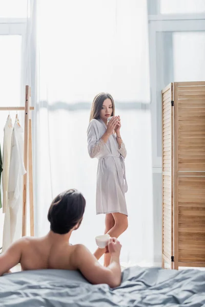 Giovane donna in abito di seta in piedi con coppa vicino a camicia fidanzato in camera da letto — Foto stock