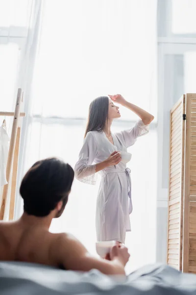 Joven mujer en túnica de seda posando con la taza cerca de novio sin camisa en el dormitorio - foto de stock