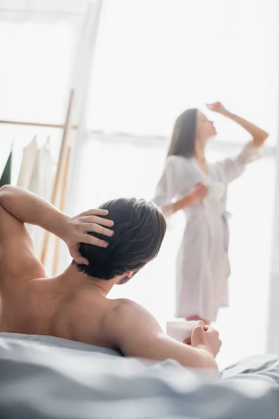 Вид сзади мужчины, смотрящего на размытую молодую женщину в шелковом халате, позирующую в спальне — стоковое фото