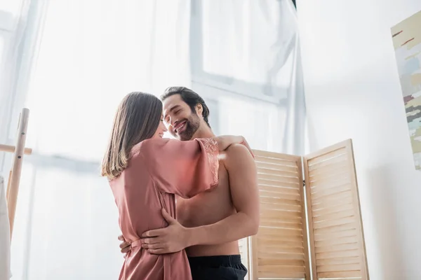 Homem barbudo e alegre abraçando namorada feliz em robe de seda rosa — Fotografia de Stock