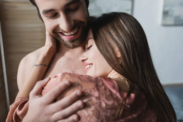 Barbudo y feliz hombre abrazando tatuado novia en rosa bata de seda - foto de stock