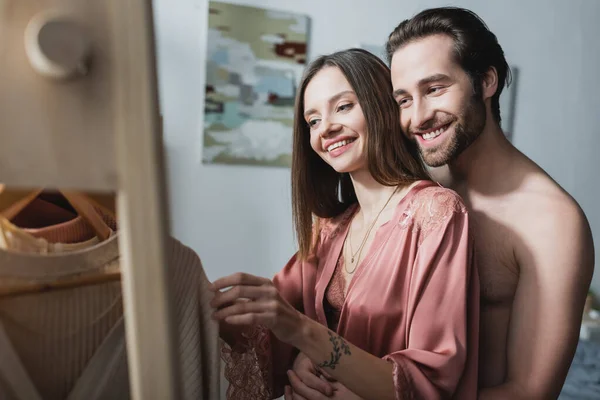 Hombre sin camisa y feliz abrazando novia tatuada en bata de seda rosa elegir ropa - foto de stock