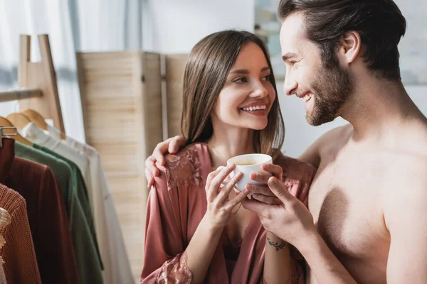 Бородатый и счастливый мужчина обнимает улыбающуюся девушку в розовом шелковом халате, держа чашку кофе — стоковое фото