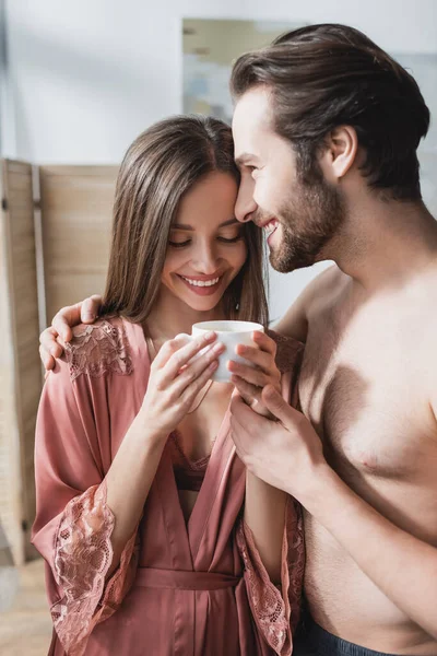 Feliz hombre abrazando sonriente novia en rosa bata de seda sosteniendo taza de café - foto de stock