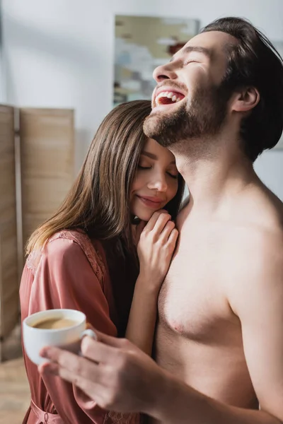 Glücklicher Mann lacht neben lächelnder Freundin in rosa Seidenmantel und hält Tasse Kaffee — Stockfoto